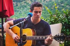 Man in grey shirt with black hair sitting in chair near red umbrella playing guitar in front of plants at Arabella Hotel Sedona in Sedona, AZ