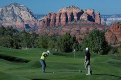 Two men playing golf on greens with one swinging golf club toward flag with red rocks in background near Arabella Hotel Sedona in Sedona, AZ