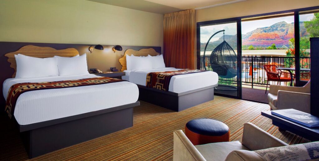 Bedroom with two beds with a chair and an outdoor patio with furniture overlooking the mountains at the Arabella Hotel in Sedona, AZ.