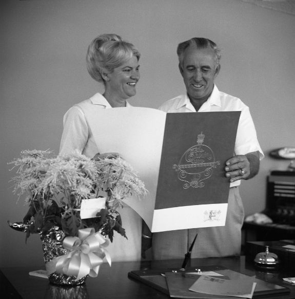 Black and white photo of two people holding booklet and smiling over table with bell, book, and a pot of flowers on it at Arabella Hotel Sedona in Sedona, AZ