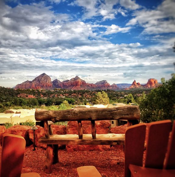 Wooden bench and chairs with blue sky filled with clouds in front of forest and mountains in background near Arabella Hotel Sedona in Sedona, AZ