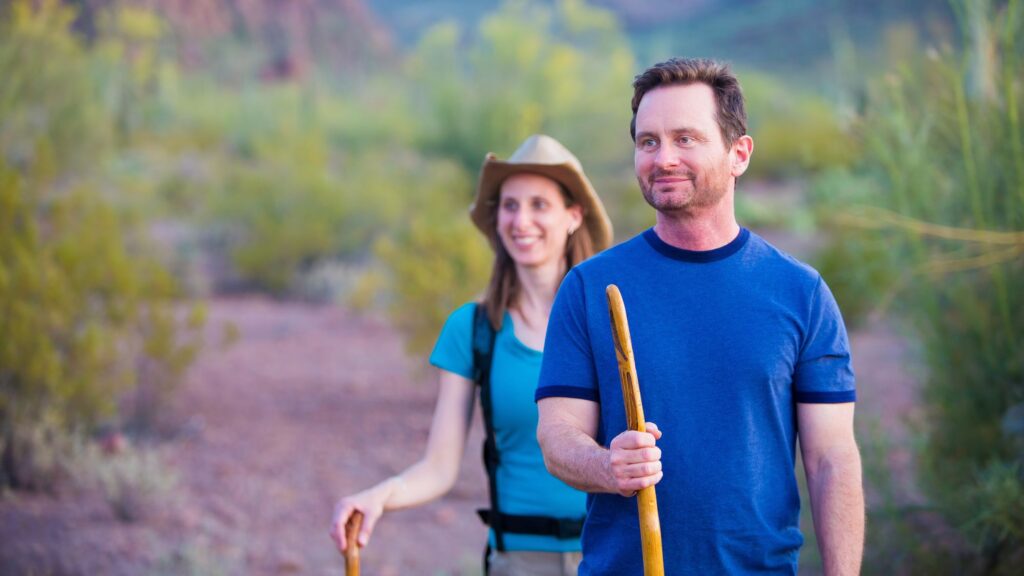 Man and woman hiking with hiking sticks in front of green foliage with mountain in background near Arabella Hotel Sedona in Sedona, AZ