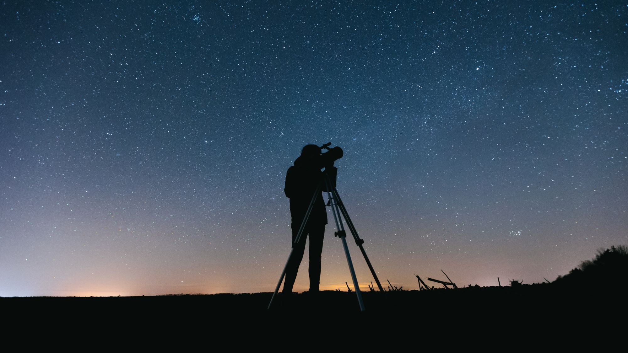 Silhouette of a person using a telescope under a starry night sky near Arabella Sedona, with a faint glow on the horizon.