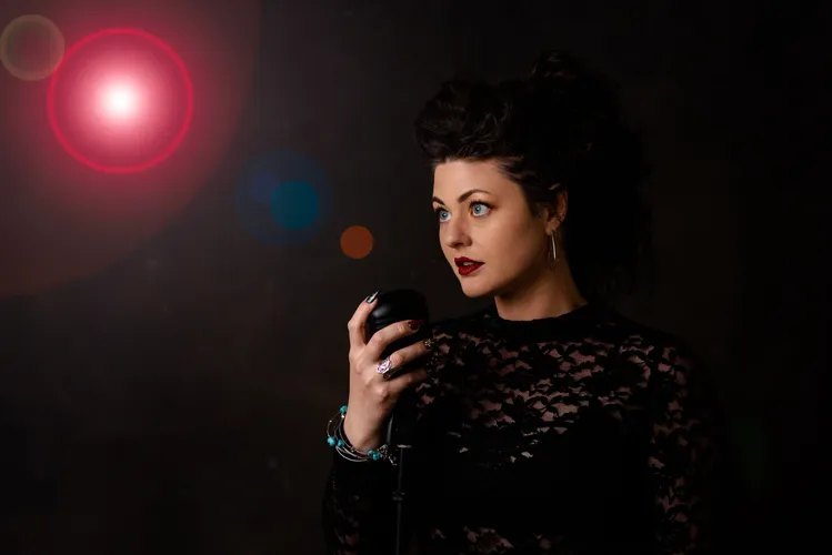 At Arabella Sedona, a woman with dark hair and red lipstick holds a vintage microphone, standing against a dark background with a bright lens flare effect.