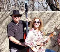 Two people smile and play upright bass and ukulele outside Arabella Sedona hotel, both wearing sunglasses in front of a stucco wall.