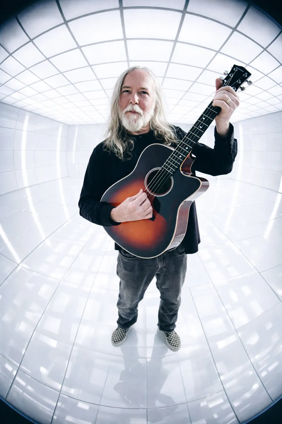 An older man with long gray hair and beard holds an acoustic guitar in a futuristic white, tiled room at Arabella Sedona, beneath bright ceiling lights.