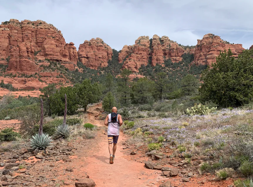 A person runs on a dirt trail near Arabella Sedona, surrounded by red rock formations and scattered greenery under a cloudy desert sky.