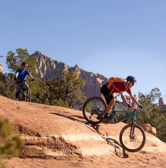 Two men mountain biking on rock surface near trees with red rock mountains in background near Arabella Hotel Sedona in Sedona, AZ