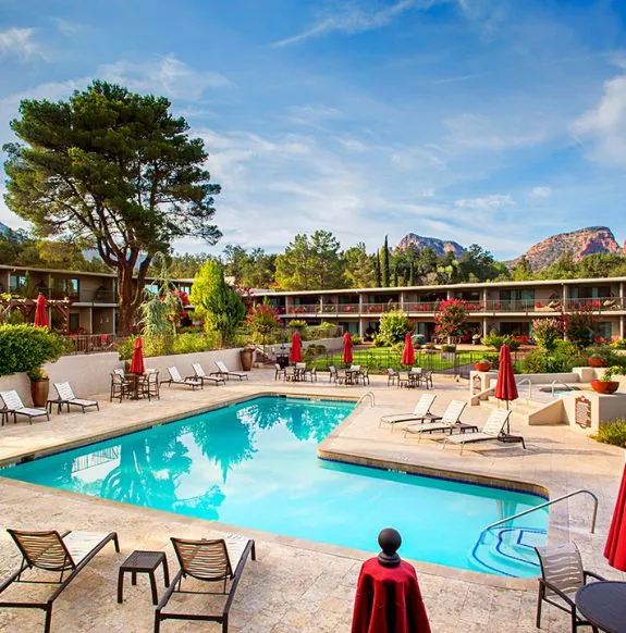 The Arabella Sedona hotel pool area with lounge chairs, red umbrellas, and lush greenery set before stunning mountains under a clear blue sky.