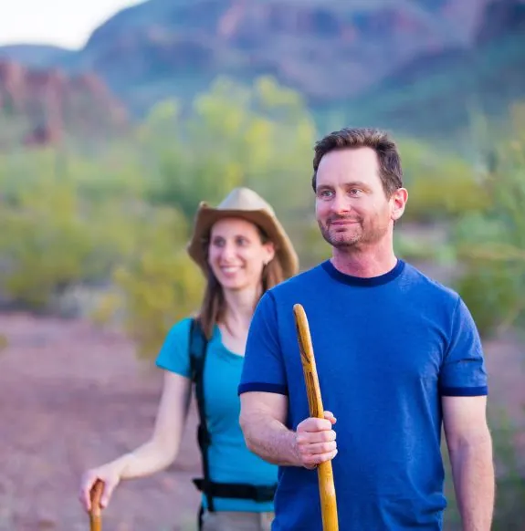 Man and woman hiking with hiking sticks in front of green foliage with mountain in background near Arabella Hotel Sedona in Sedona, AZ
