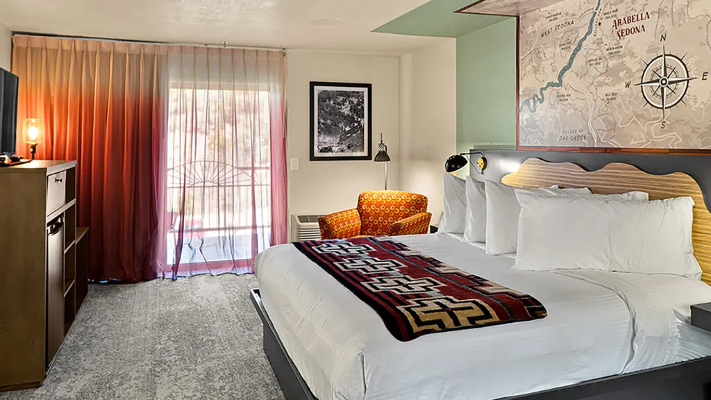 A hotel room with a large bed, a window with red curtains, a chair, a TV, and a map on the wall.