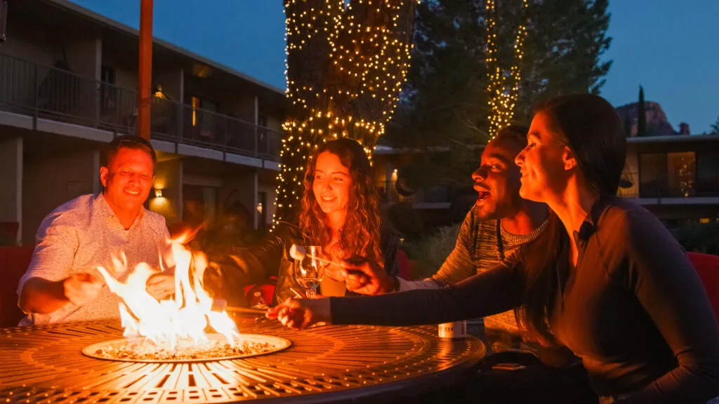 Four people relax by a fire pit at night at Arabella Sedona, smiling and talking as string lights on trees create a warm, festive atmosphere.