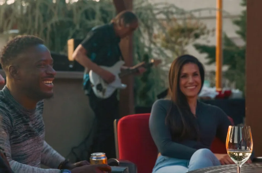 Two people smile while sitting outdoors at Arabella Sedona; a guitarist plays in the background, and a glass of white wine sits on a table in the foreground.