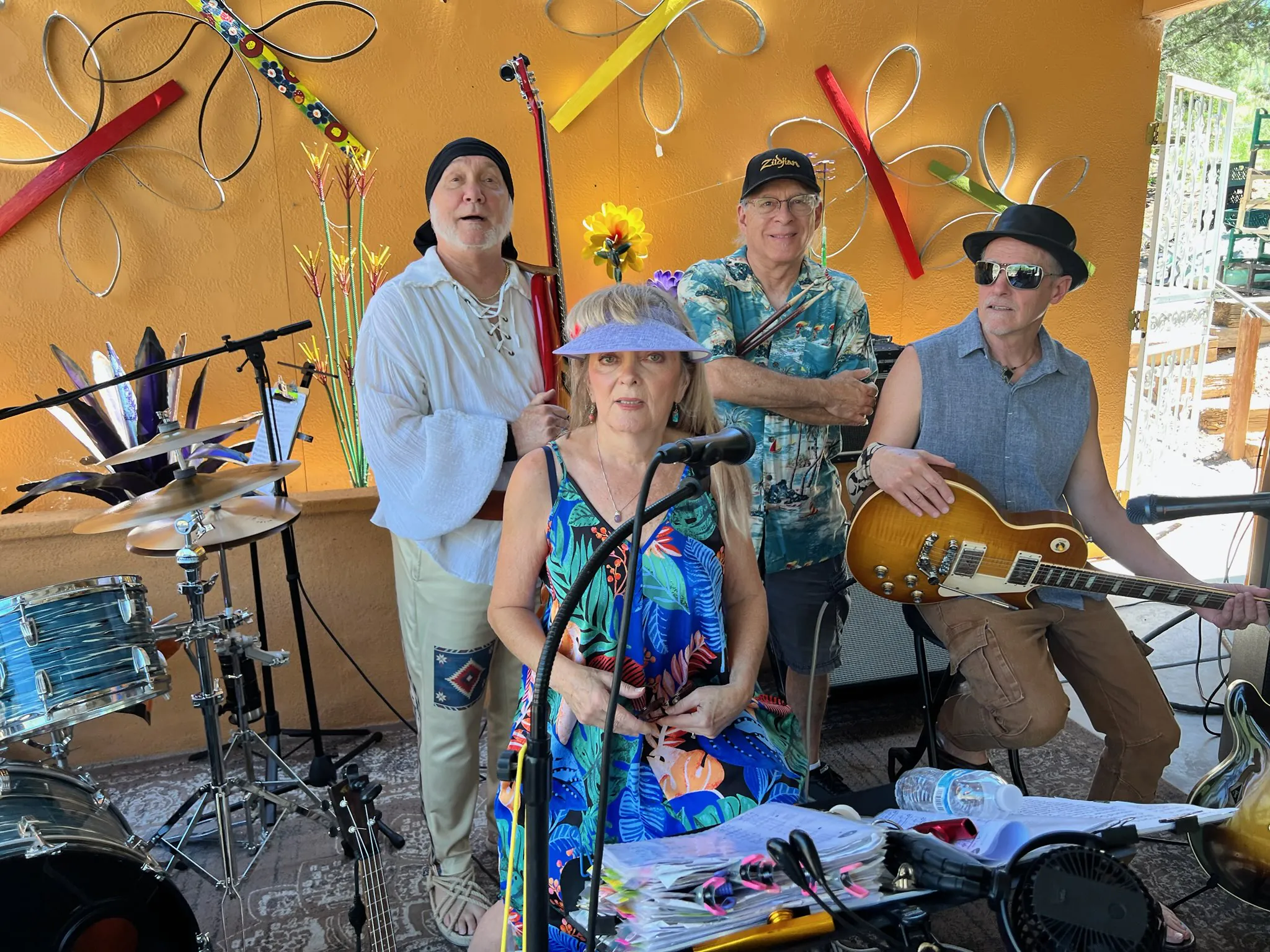 At Arabella Sedona, four musicians pose with instruments outdoors by colorful wall art; the front woman wears a sunhat and floral dress.