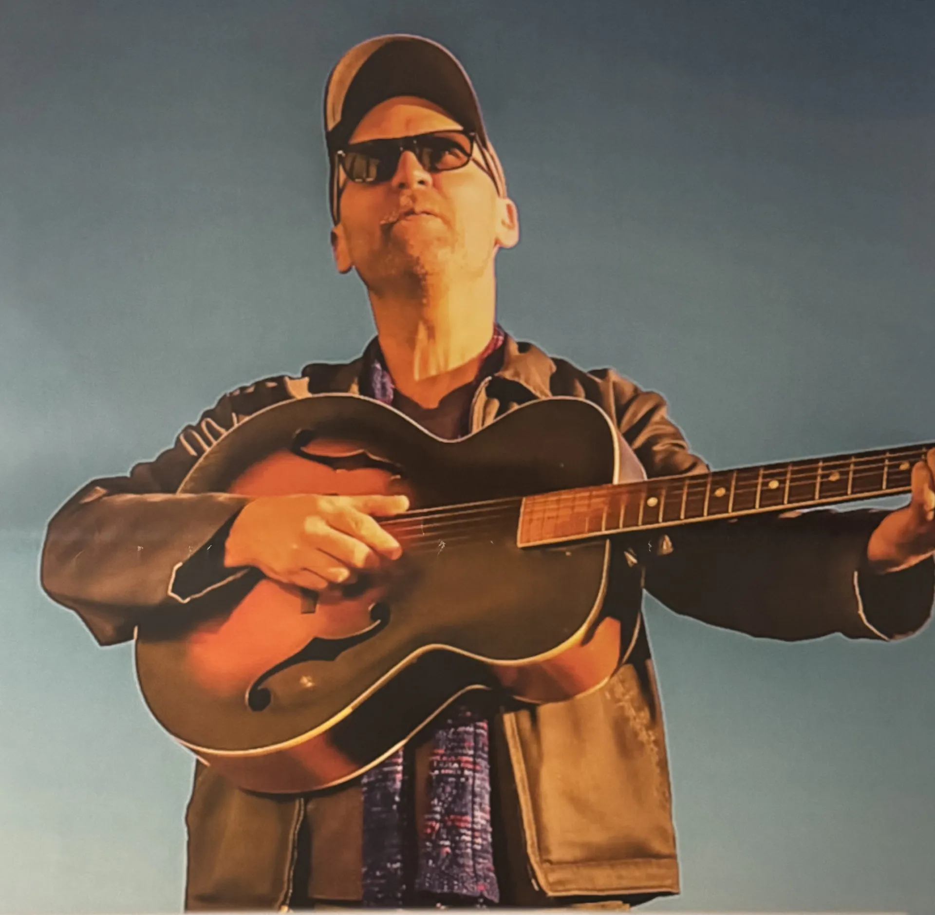 Man in sunglasses and a cap plays an acoustic guitar outdoors under a clear blue sky at Arabella Sedona.